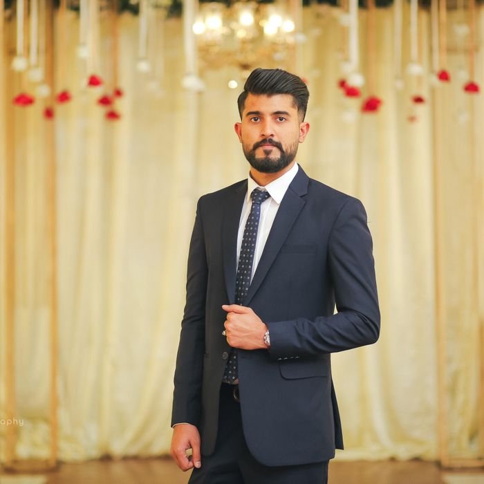 Bearded man in a dark suit poses confidently in a formal indoor setting.
