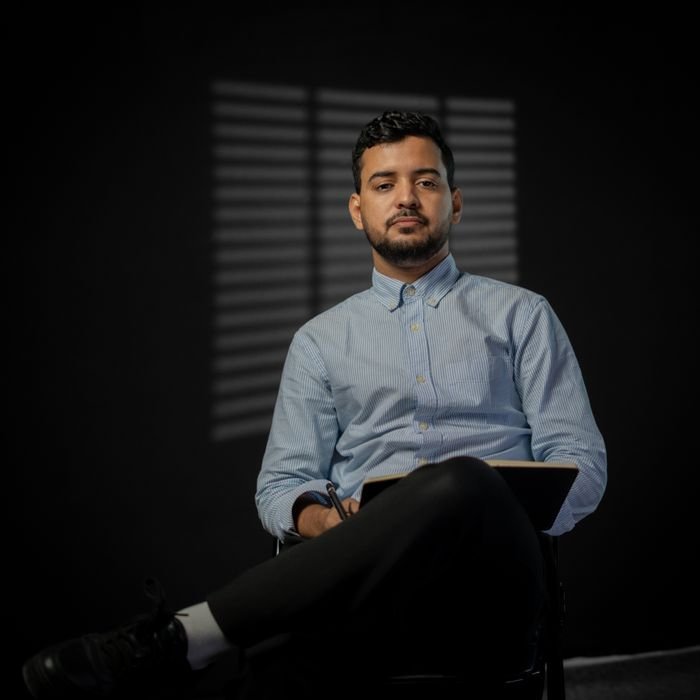 gardener-testimonial-3 Portrait of a confident businessman seated indoors in a dark, moody setting, holding a notebook.